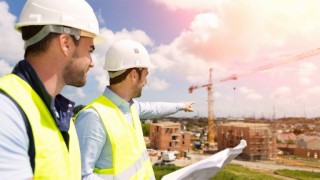 Zwei Männer mit Schutzhelmen schauen auf eine Baustelle.